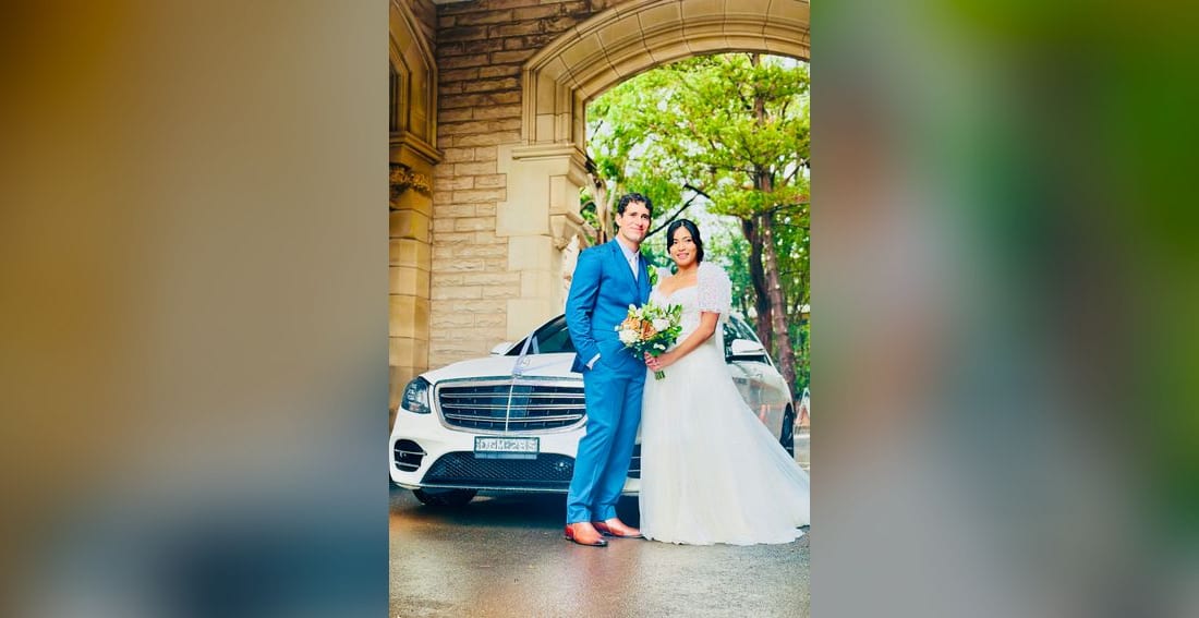 Bride and groom beside a white Mercedes S Class AMG sedan under a heritage sandstone arch