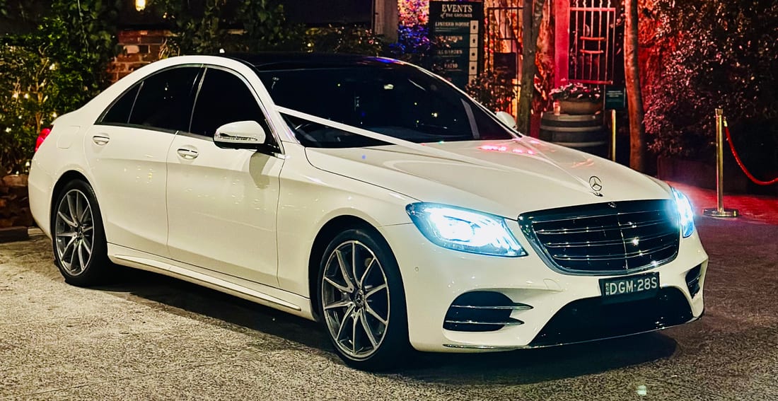 White Mercedes S Class AMG sedan, front three-quarter with headlights on, at an event venue at night