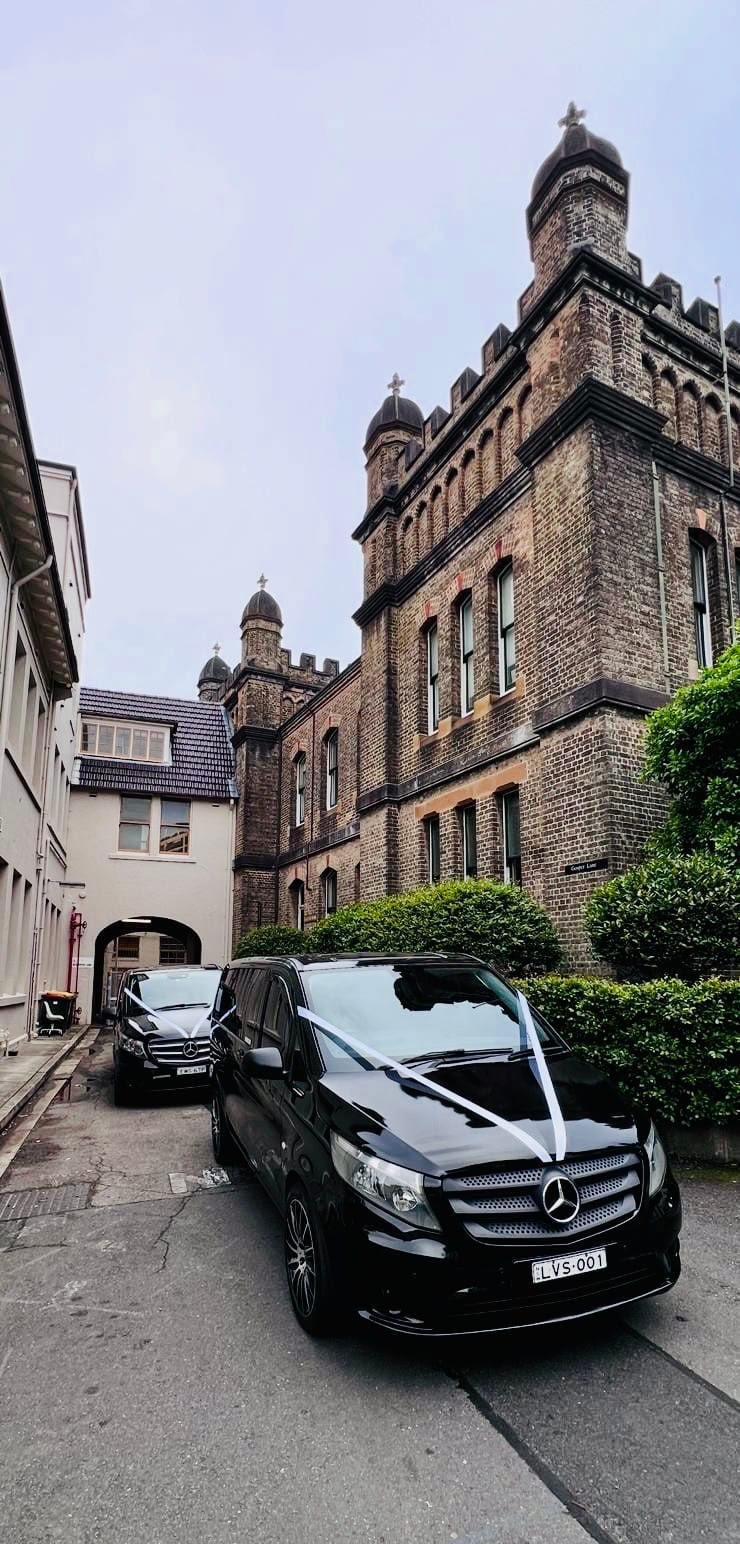 Two black Mercedes Valente wedding minivans with ribbons parked beside a heritage sandstone building