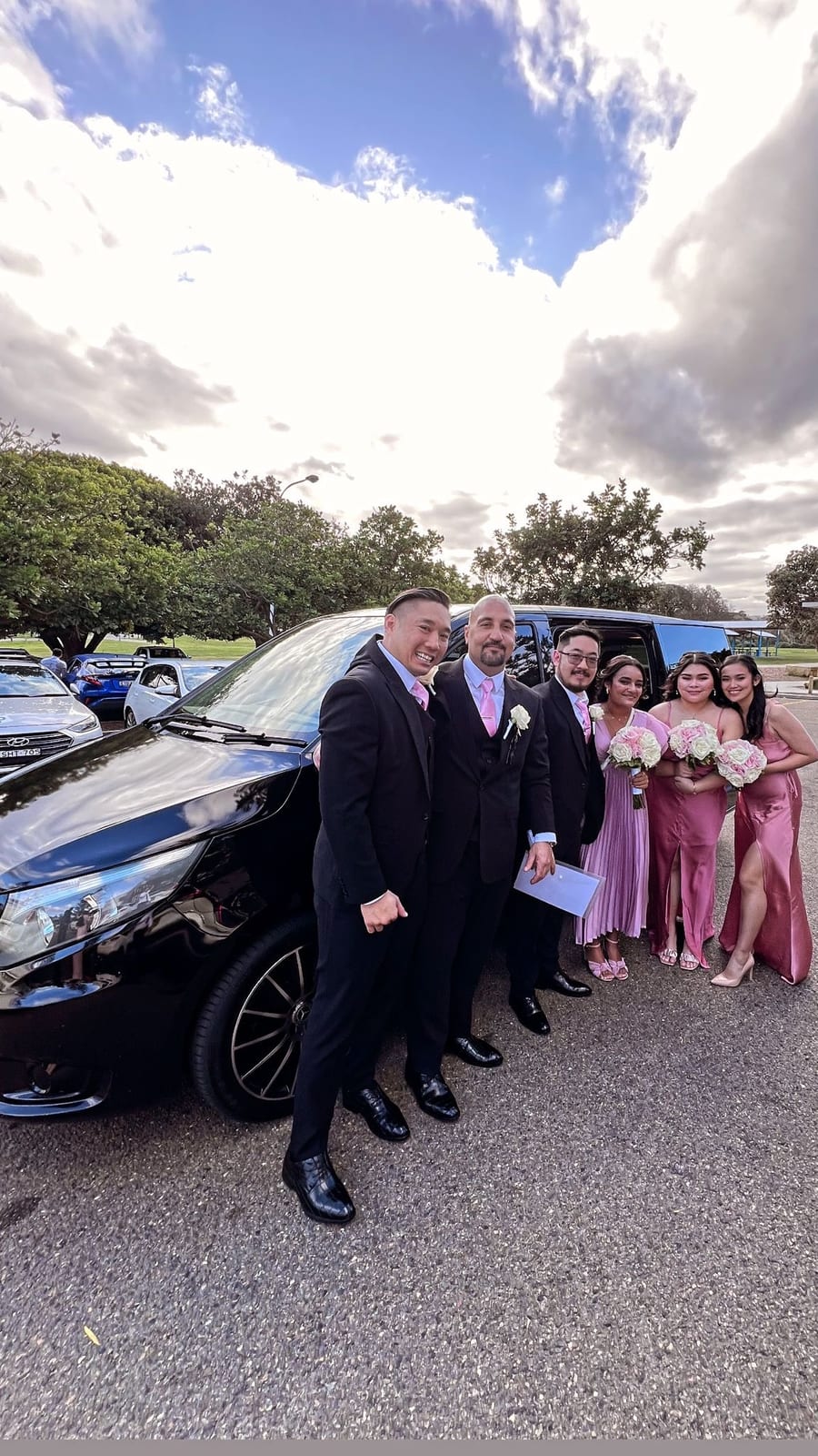 Wedding party in suits and pink gowns beside a black Mercedes Valente minivan